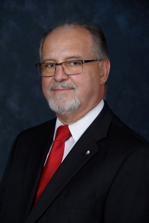 Headshot of Edmund Zielinski, a white male with gray hair and a beard; he wears a black suit jacket, a white button-up, and a red tie. 