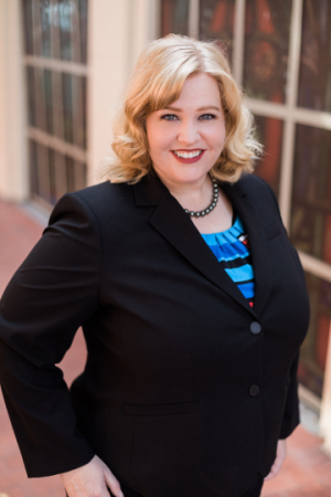 Headshot of Andrea Townsend, a white female with blonde hair; she wears a black suit jacket over a blue and black horizontal striped blouse. 